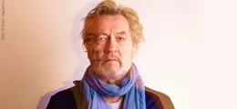 Ein Mann mit grauem Haar und Bart, trägt einen blauen Schal, schaut direkt.