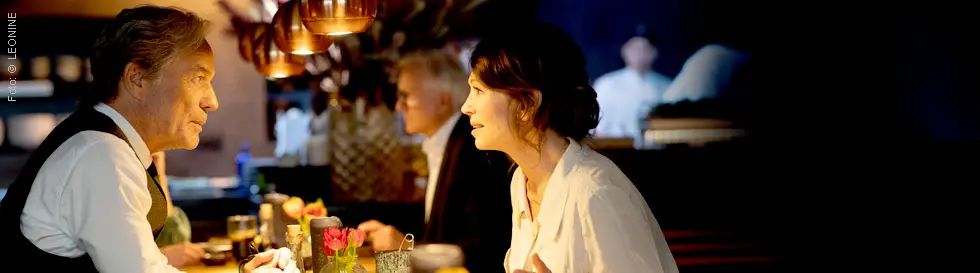 Eine Frau und ein Mann unterhalten sich an einem Tisch, im Hintergrund verschwommene Gestalten.