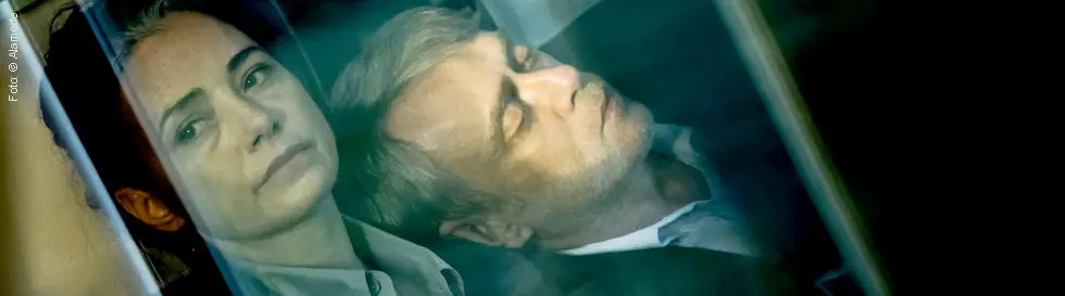 Zwei Personen sitzen eng beieinander, eine Frau schaut nachdenklich, während ein Mann mit geschlossenen Augen lehnt.