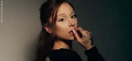 Eine Frau mit dunklem Haar, Tattoos auf der Hand, berührt ihre Lippe nachdenklich, erschocken.