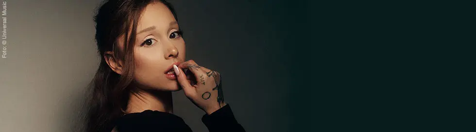 Eine Frau mit dunklem Haar, Tattoos auf der Hand, berührt ihre Lippe nachdenklich, erschocken.