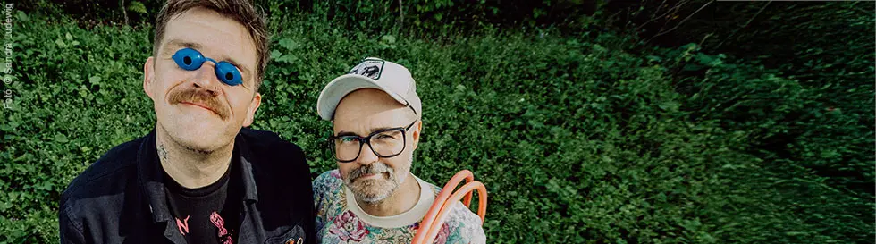 Zwei Männer, einer mit blauer Brille und Schnurrbart, der andere mit Mütze und Brille, stehen auf einer grünen Wiese.