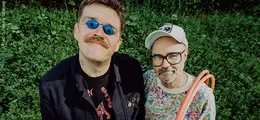 Zwei Männer, einer mit blauer Brille und Schnurrbart, der andere mit Mütze und Brille, stehen auf einer grünen Wiese.