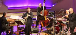 Männer in Anzügen spielen Saxophon, Flöte, Klavier, Bass und Schlagzeug auf einer Bühne mit lila beleuchtetem Hintergrund.