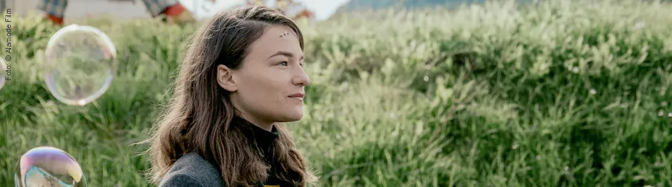 Eine Frau mit langen Haaren im Profil vor einer grünen Wiese, Seifenblasen umgeben sie.