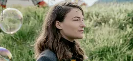Eine Frau mit langen Haaren im Profil vor einer grünen Wiese, Seifenblasen umgeben sie.