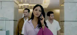 Eine Frau in lila Bluse telefoniert mit ernstem Gesichtsausdruck, zwei Männer im Hintergrund. Elegante, moderne Umgebung.