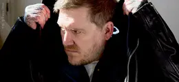 Ein Mann zieht den Kragen seiner schwarzen Jacke hoch. Er hat tätowierte Hände und kurze Haare.