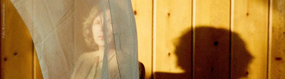 Ein Vorhang verdeckt teilweise eine Person, deren Gesicht verschwommen hinter dem Stoff zu erkennen ist; Holzvertäfelung im Hintergrund.