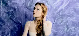 Eine Frau mit langen, roten Haaren blickt nach oben. Der Hintergrund ist in lila Tönen gestaltet.
