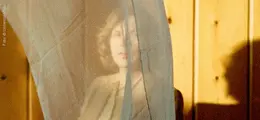 Ein Vorhang verdeckt teilweise eine Person, deren Gesicht verschwommen hinter dem Stoff zu erkennen ist; Holzvertäfelung im Hintergrund.