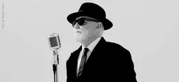 Ein Mann mit Hut und Sonnenbrille steht vor einem Retro-Mikrofon, in einem schwarz-weißen Setting.