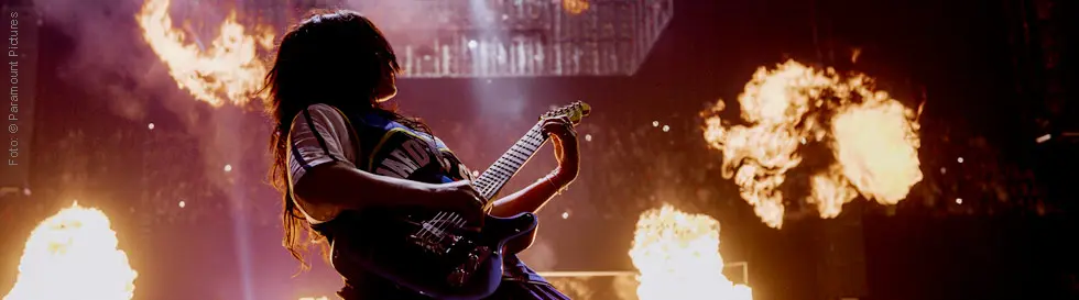 Eine Person kniet auf der Bühne und spielt Gitarre, umgeben von Flammen im Hintergrund.