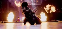 Eine Person kniet auf der Bühne und spielt Gitarre, umgeben von Flammen im Hintergrund.