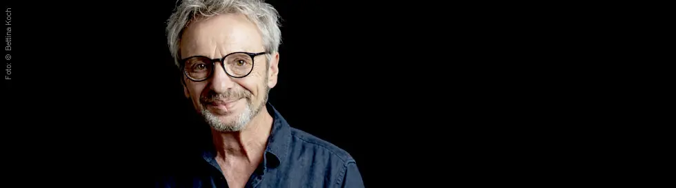 Ein lächelnder Mann mit Brille und grauem Haar vor schwarzem Hintergrund.