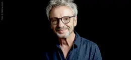 Ein lächelnder Mann mit Brille und grauem Haar vor schwarzem Hintergrund.