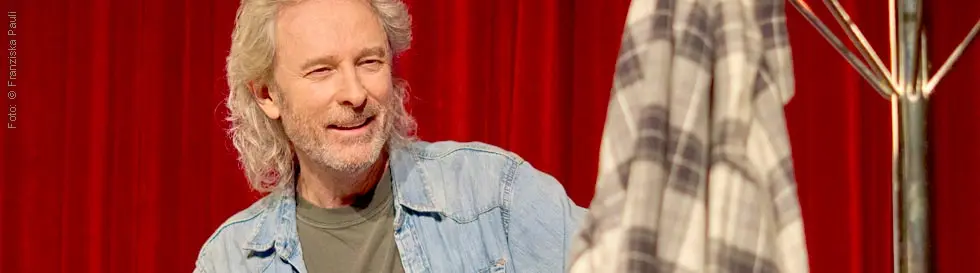 Ein Mann mit grauem Haar in Jeanshemd vor rotem Vorhang hält kariertes Tuch.