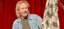 Ein Mann mit grauem Haar in Jeanshemd vor rotem Vorhang hält kariertes Tuch.