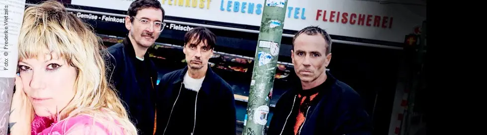 Vier Personen stehen vor einem Laden, eine lehnt lässig an einem Pfeiler.