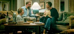 Vier Menschen sitzen in einem gemütlichen Wohnzimmer und unterhalten sich lachend. Im Hintergrund steht eine Lampe.