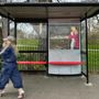 Ein Wartehäuschen mit einer Frau im Vordergrund. Klare Struktur, leuchtend roter Sitz. Hintergund: Park und Bäume., CD