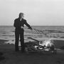 Eine Person steht am Strand, hält ein langes Objekt und bewegt es über ein knisterndes Feuer. Im Hintergrund das Meer., CD