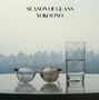 Text: "SEASON OF GLASS YOKO ONO". Eine Brille und ein halbvolles Glas Wasser stehen auf einem Tisch mit Stadt im Hintergrund.
