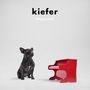 Text: "kiefer happysad". Ein schwarzer Hund sitzt neben einem kleinen roten Spielzeugklavier auf hellem Hintergrund.