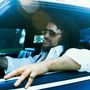 Ein Mann mit Hut und Sonnenbrille lehnt lässig im Auto, eine Zigarette im Mund., MC
