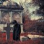 Ein Mann in einem dunklen Mantel steht nachdenklich vor einem alten Grabstein auf einem rot gefärbten Friedhof.