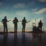 Silhouetten von vier Musikern: drei mit Gitarren, einer am Schlagzeug, vor einem Himmel mit Wolken im Hintergrund.