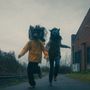 Zwei Personen laufen Hand in Hand, tragen Tiermasken und Jacken, links gelb, rechts braun, in industrieller Umgebung., CD