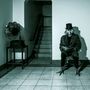 Ein Raum mit Schallplattenspieler, Treppe im Hintergrund. Person mit Vogelmaske und Zylinder sitzt lesend auf einem Stuhl., LP