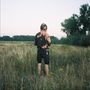 Eine Person mit gesprenkeltem Hoodie steht in einem Feld. Hohe Gräser und Bäume im Hintergrund., LP