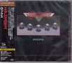 "AEROSMITH" oben in Rot, "ROCKS" unten in Weiß. Fünf leuchtende Edelsteine auf schwarzem Hintergrund. Japanische Schrift links.