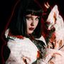 Eine Frau mit schwarzem Haar und dramatischem Make-up, in einem strukturierten weißen Mantel, vor einem rot beleuchteten Hintergrund., LP