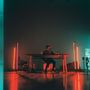 Kein Text vorhanden. Szene: Person an Tisch mit Laptop, umgeben von buntem Licht. Technik und Kabel sind sichtbar., CD