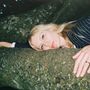 Eine Person mit blonden Haaren liegt auf einem moosigen Baumstamm und schaut zur Seite. Sie trägt einen Anzug mit Streifen., LP