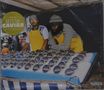 "Bitume Caviar" steht auf einer gelben Tafel. Zwei Männer in gestreifter Kleidung und gelben Schürzen stehen am Marktstand mit Konserven., CD