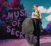 "MUSIC OF SECRETS" steht in lila Buchstaben. Eine Person tritt in die Kamera, Hintergrund aus lila Bäumen und blauer Himmel., CD