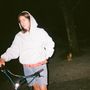 Eine Frau mit Kapuzenpulli hält ein Fahrrad in der Hand, im Hintergrund sitzt eine Katze auf dem Boden., LP
