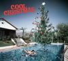 Text "Cool Christmas". Eine Frau schwimmt im Pool mit Weihnachtsbaum. Haus und Palmen im Hintergrund.