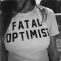 Auf einem T-Shirt steht fett "FATAL OPTIMIST". Eine Person trägt es, im Freien, mit langen Haaren. Schwarz-Weiß-Foto., LP