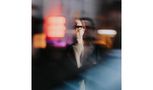 Ein unscharfes Farbfoto zeigt das verschwommene Gesicht eines Mannes mit Brille hinter einer Scheibe. Neonlichter im Hintergrund., CD