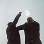 Zwei Personen in Anzügen stehen unter einem klaren Himmel und strecken die Arme zum Mond empor., LP