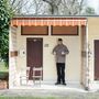 Eine Person in einer Veranda mit Daumen hoch, neben zwei Stühlen, einer Tür mit Nummer "591" und einem orange-gestreiften Vordach., LP