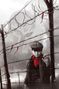 Stacheldrahtzaun, Junge mit rotem Hemd, neblig, Schneeflocken fallen, düstere Atmosphäre, große Maske im Hintergrund.