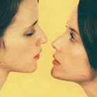 Zwei Personen stehen sich nah gegenüber vor einem gelben Hintergrund. Sie blicken einander intensiv an.