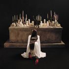 Der Text „ESTRUS“ steht auf dem Rücken einer knieenden Person vor einem Altar mit vielen Kerzen und einer schwarzen Katze.