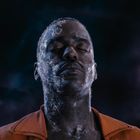 Ein Mann mit geschlossenen Augen, Eiskristalle auf Gesicht und Hals, trägt eine orange Jacke vor einem dunklen, nebulaartigen Hintergrund.